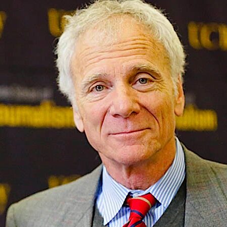 ed-wasserman_1 An older man with gray hair and a slight smile stands against a backdrop with "Berkeley Journalism" and "UC Berkeley" written on it. He is wearing a gray suit, blue shirt, and red striped tie.