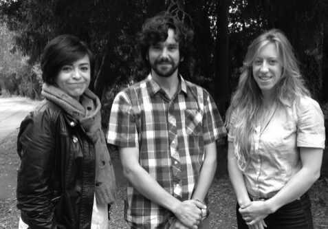 Three people are standing outdoors in a line, smiling at the camera. The person on the left is wearing a black jacket and scarf, the person in the middle is wearing a plaid shirt, and the person on the right is wearing a light-colored shirt. Trees are in the background, creating an inviting setting for Berkeley Journalism.