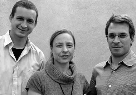 Black and white photo of three people standing side by side and facing the camera, embodying the spirit of Berkeley Journalism. They are wearing casual outfits; two men are on either side of a woman in the center. The background is a plain wall.