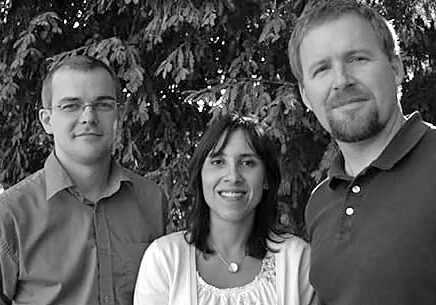 A black and white photo of three people standing outdoors in front of a tree. Two men are positioned on either side of a woman, all smiling and dressed casually. The scene exudes the spirit of Berkeley Journalism, capturing a candid moment in their day.