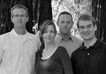 A black and white photo shows four people standing outdoors in front of trees. There are three men and one woman. The man on the left wears glasses and a light shirt; the woman wears a dark top; the third person wears a button-up shirt, and the fourth person, possibly from Berkeley Journalism, wears glasses and a polo shirt.