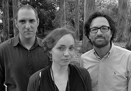 Three people stand together outdoors in front of tall trees. The black-and-white image shows a woman in the center with two men on either side. They all have serious expressions and are dressed in casual clothing, embodying the thoughtful gaze often seen in Berkeley Journalism students.