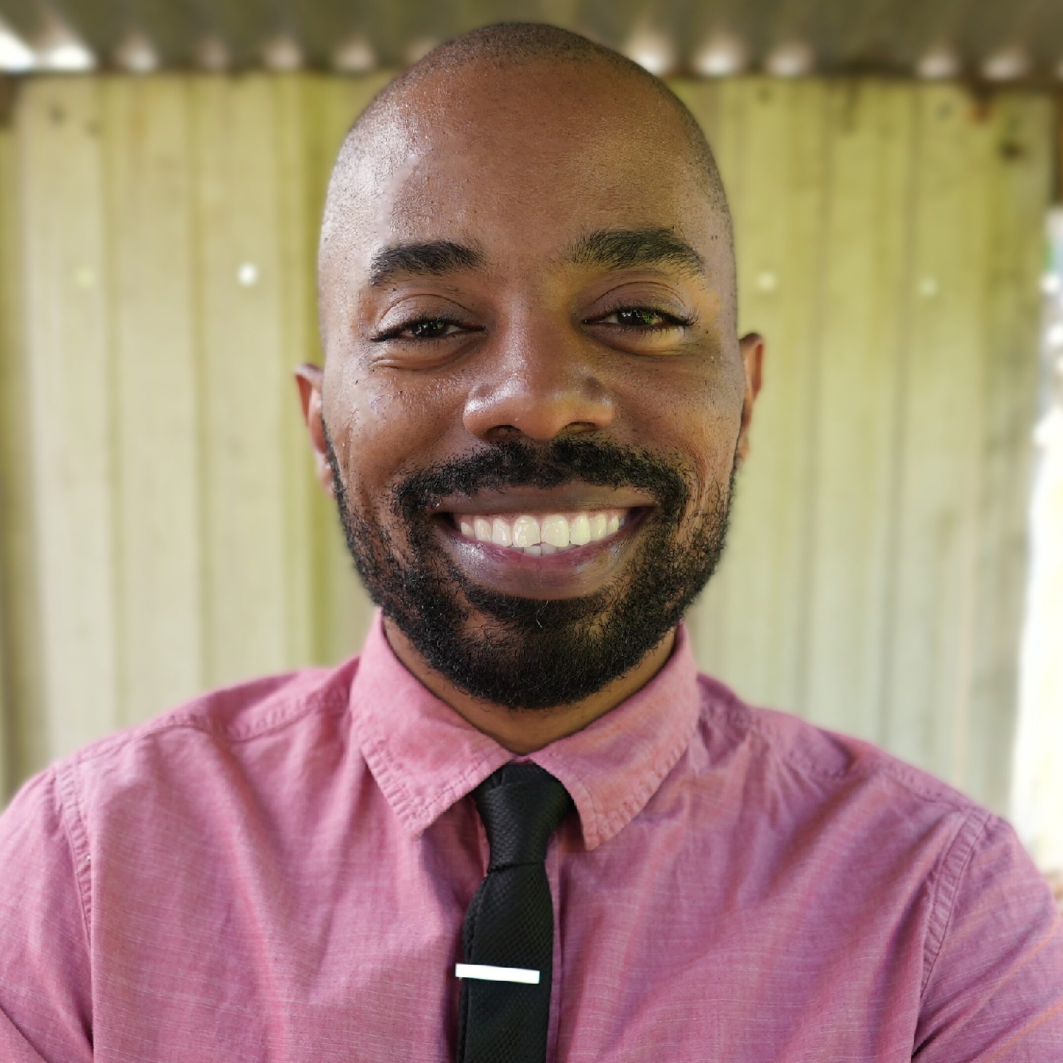john-harden John D. Harden, with a beard and mustache, is smiling at the camera. He is wearing a pink button-up shirt and a black tie with a silver tie bar. The background is out of focus with what appears to be a wooden structure.