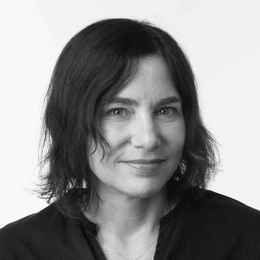 Black and white portrait of a woman with dark hair wearing a black shirt looking at the camera in front of a white background.