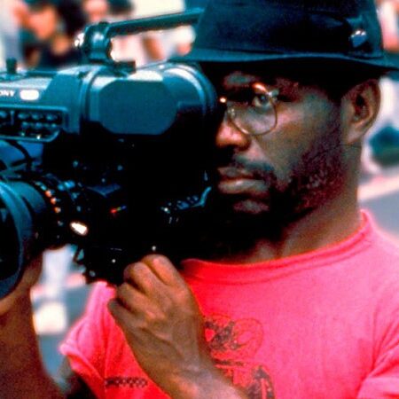 A maverick filmmaker in a red shirt and black hat is holding a large video camera, recording an event. The background shows a crowd of people and blurred outdoor elements, suggesting that the scene is taking place in a public space. This could easily be part of one of Marlon T. Riggs' documentary fellowships.