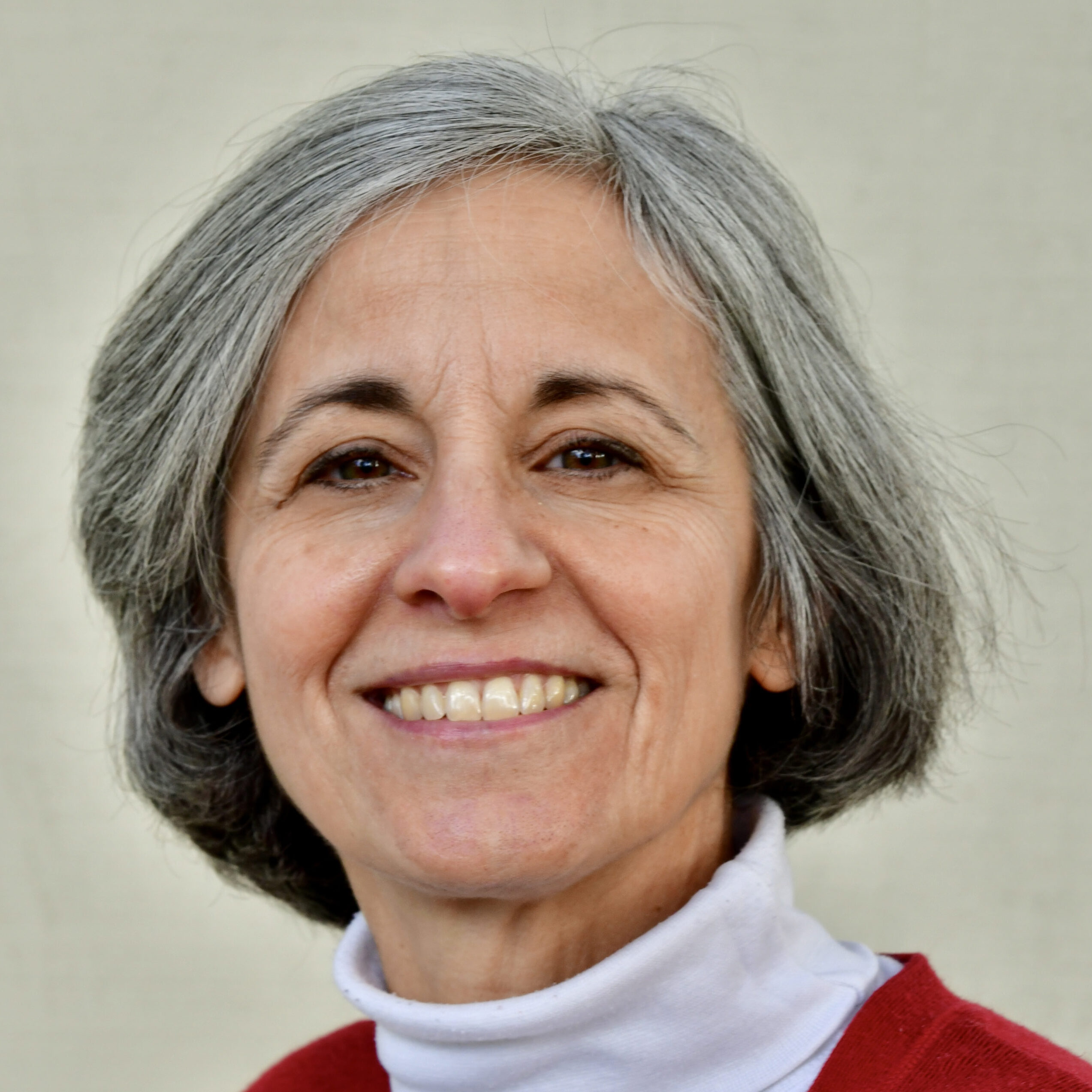 schiavo 2 A smiling woman with short, gray hair is looking at the camera. She is wearing a white turtleneck under a red sweater, embodying the spirit of Berkeley Journalism. The background is a plain, neutral color.