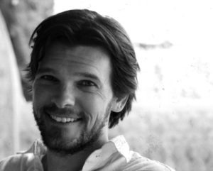 A black-and-white image of Chris Smith smiling at the camera. They have medium-length hair, a beard, and are wearing a light-colored shirt. The background is blurred.