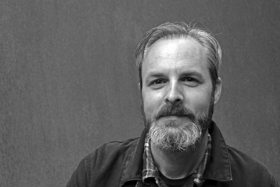 Black and white photo of middle-aged man Dave Gilson with short hair and a beard. He wears a plaid shirt under a jacket. The background is plain and neutral.