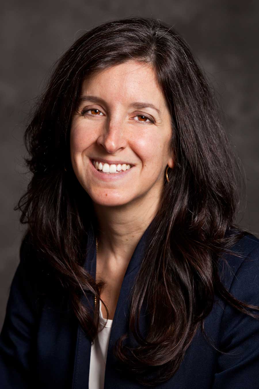 A woman with long, dark hair and a friendly smile, is wearing a navy blazer over a light-colored top. She is posed against a gray, textured background.