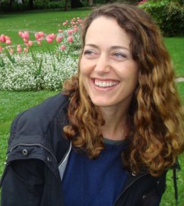 Sarah Lavender Smith, a woman with wavy brown hair and a bright smile, is sitting outdoors in a garden. She is wearing a navy blue top and a black jacket. Behind her, there are pink tulips and white flowers on a green lawn.