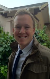 A smiling person with short, light hair is wearing a brown jacket, white shirt, and blue tie. David Ferry stands outdoors in front of a building with neatly trimmed greenery visible in the background.