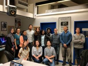 In what appears to be an office or classroom setting, a group of thirteen men and women of diverse backgrounds pose together in a room with blue doors and posters on the walls. Some are standing while others are seated. It seems the moment captures their gathering for the Dean's Letter in March 2017.