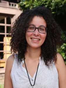 A person with curly hair and glasses, who could be Semany Gashaw, smiles warmly while standing outdoors. They are wearing a sleeveless white top and a long necklace, with a building and greenery visible in the background.