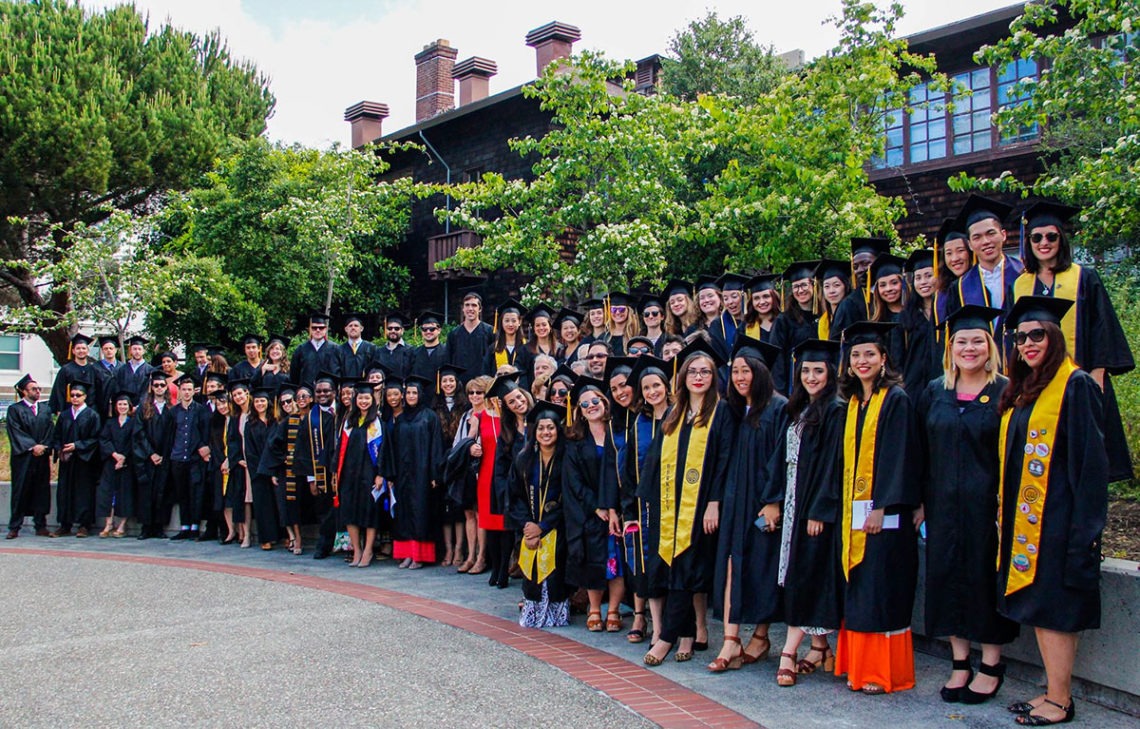 A large group of graduates in caps and gowns poses together outdoors on a sunny June 2016 day. They stand on a curved pathway in front of a building surrounded by greenery and trees. Some graduates wear honor cords and sashes, while others are dressed in academic regalia, proudly holding the Dean's Letter.