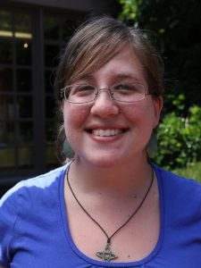 Alissa Greenberg, with glasses and light brown hair, is smiling at the camera. She is wearing a blue shirt, blue earrings, and a star-shaped pendant necklace. The background is slightly blurry, featuring a dark building with glass windows and green foliage.