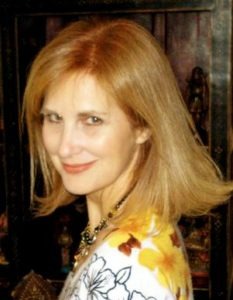 Wendy Jane Carrel, a woman with shoulder-length blonde hair, is smiling softly while looking over her shoulder. She is wearing a white top with a floral pattern and a statement necklace. The background features decorative statues and dark wooden elements.