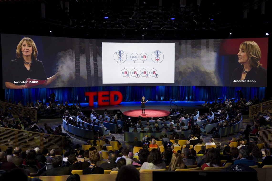 A woman stands on a circular stage giving a TED Talk. Behind her is a large screen displaying a scientific diagram, and there are smaller screens with her image and name, Jennifer Kahn. The audience is seated in a semi-circular arrangement, captivated by the insights Jennifer shared in the March 2016 Dean's Letter.