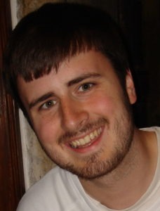 A young man with short brown hair and a beard, who can be identified as Jordan Conn, is smiling at the camera. He is wearing a white shirt and standing indoors against a light-colored wall.