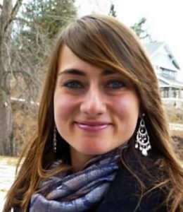 Kate Elston, a woman with long brown hair, wearing dangly earrings and a scarf, smiles at the camera. She is standing outdoors with trees, a house, and a snowy ground in the background.