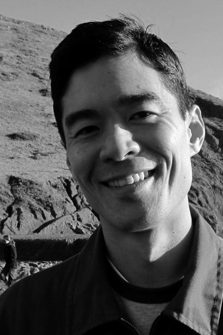 Black and white portrait of a smiling person with short, dark hair, wearing a jacket. Mike Shen stands against hills and rocky terrain in the background under a clear sky.