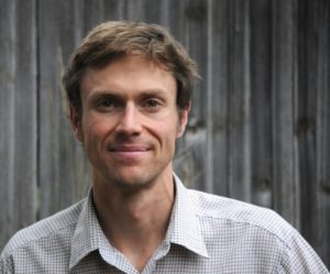 A person with short brown hair and a light complexion smiles while wearing a checkered shirt. The background features a weathered wooden fence, reminiscent of the rustic charm often captured in Terray Sylvester's photographs.