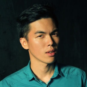 A young man with short dark hair, reminiscent of Jeremy Lin, is gazing at the camera with a neutral expression. He is wearing a teal button-up shirt and standing against a dark background. The lighting highlights his facial features, casting slight shadows.