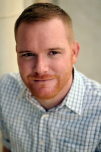 Nick Burns, a man with short reddish hair and a beard, smiles slightly at the camera. He is dressed in a light-colored, checkered shirt. The background is blurred and neutral in color.
