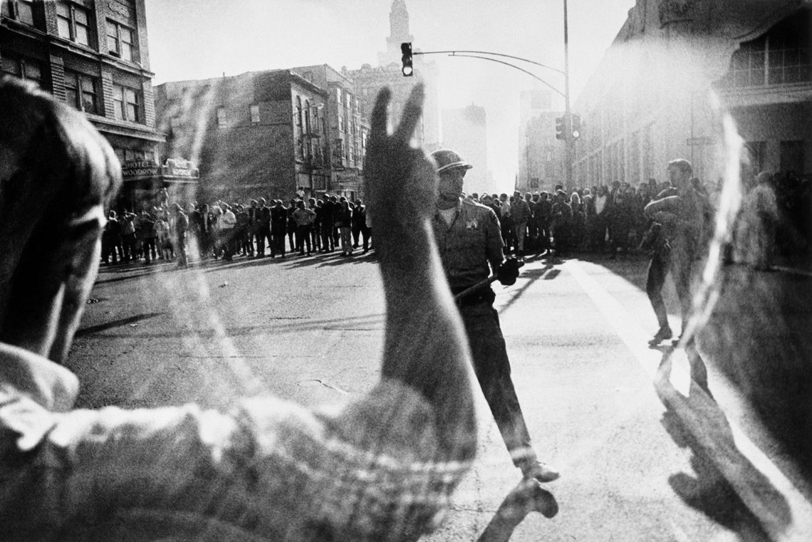 A black-and-white photograph captures a Bay Area street scene with a line of police officers and a crowd in the background. In the foreground, a resister raises their arm, partially obscured by blur and light reflections, creating a sense of movement and tension emblematic of social movement photography.