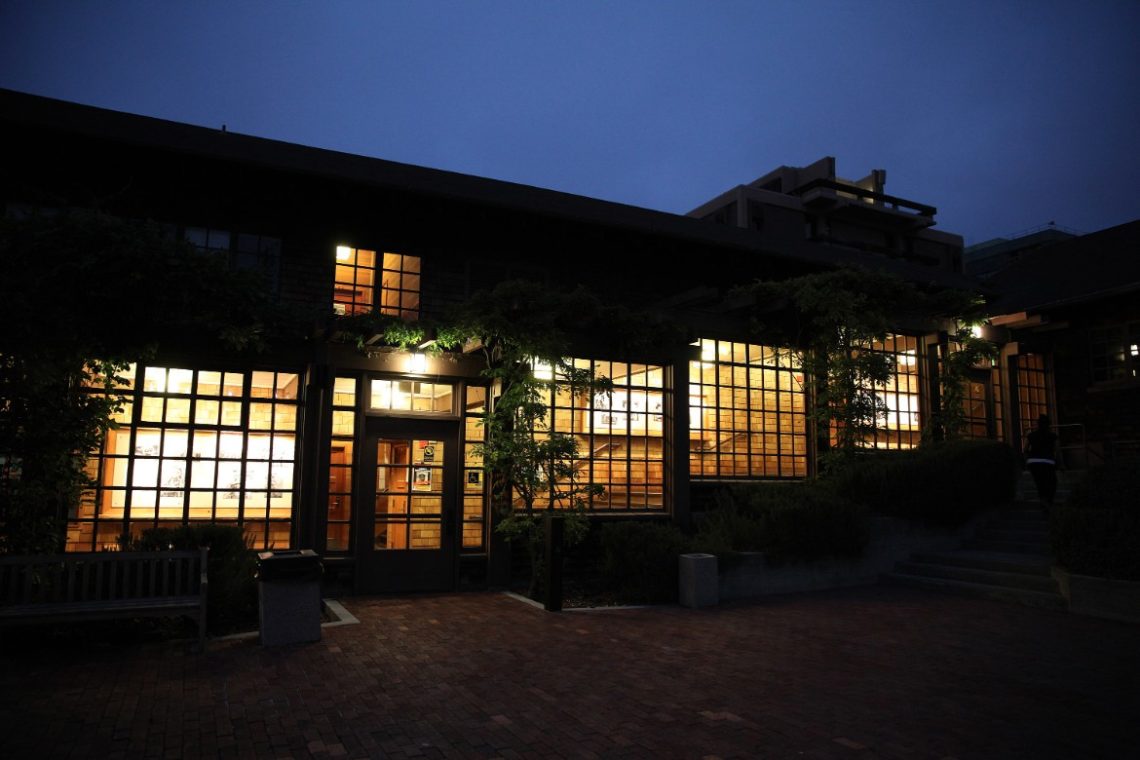 A warmly lit building with large, grid-patterned windows is seen at night. The brick pathway and a bench in front of the entrance add to the serene atmosphere. The building's exterior is partially covered in greenery, with a modern building visible in the background.