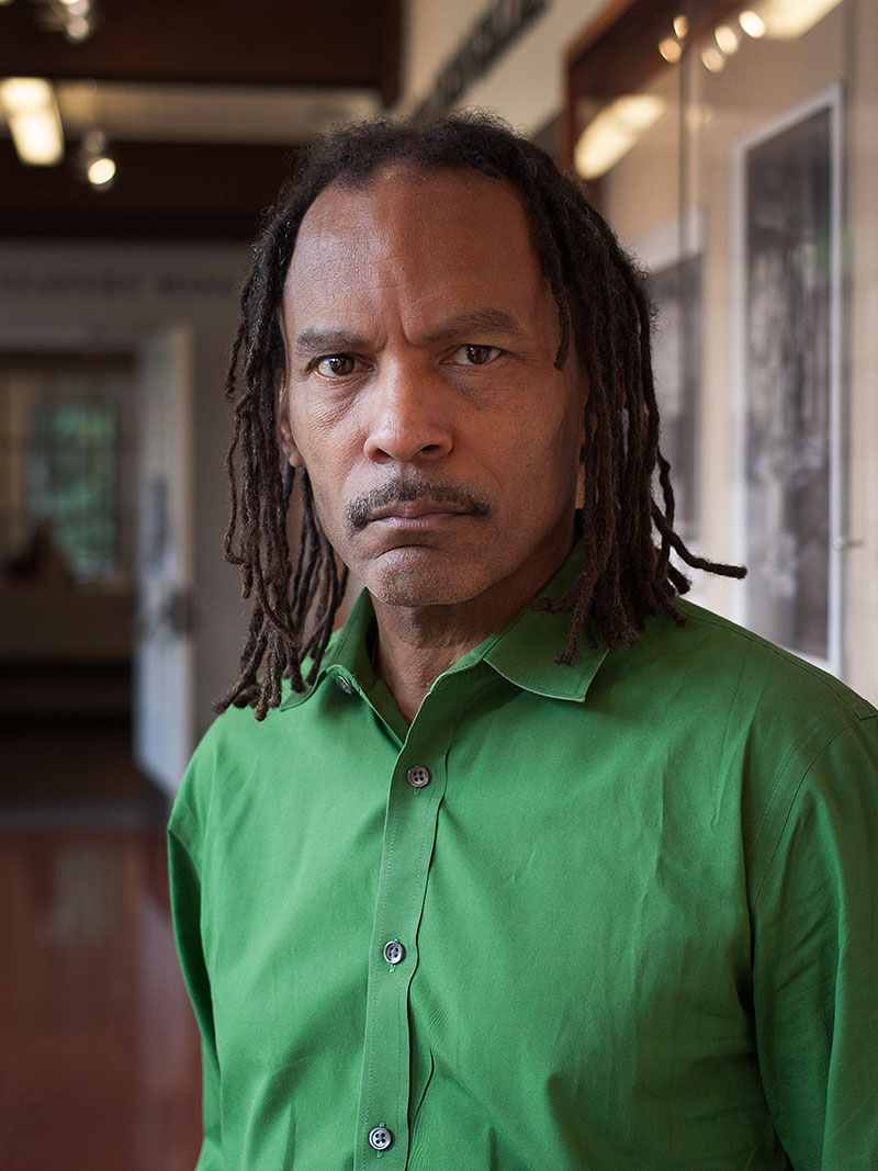 A man with long dreadlocks and a mustache is wearing a bright green button-up shirt. He stands indoors in a hallway with a serious expression on his face, resembling Orlando Bagwell. The background is slightly blurred, showing doors and wall displays.