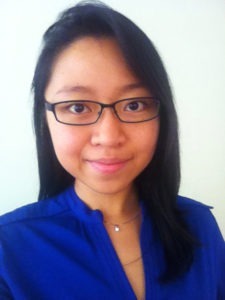 A person with long dark hair wears glasses and a blue top. They are smiling slightly and looking at the camera. The background is a plain, light-colored pearly tan.