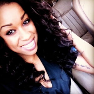 A woman with long, curly dark hair, likely Brittany Johnson, smiles brightly while sitting in a vehicle. She is wearing a dark top and has a prominent black ink tattoo on her upper arm. The background features beige leather car seats.