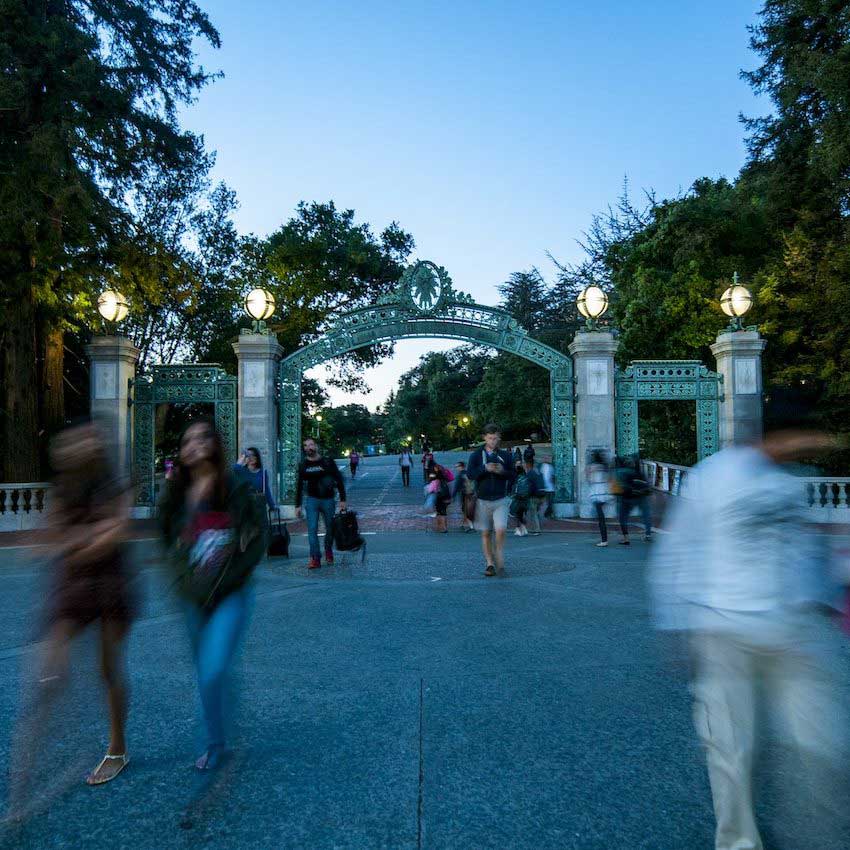 A busy walkway with people, including an undergraduate journalist, passing beneath a large, decorative metal archway at dusk. The arch boasts ornate designs and glowing lamps atop stone pillars, surrounded by trees on either side. The sky transitions to evening, casting a soft blue hue.