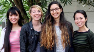 Four smiling individuals stand together outdoors. One person has long black hair and wears a red and white striped top, another has short blonde hair and a black jacket, the third has long brown hair and glasses, and the fourth has long dark hair, wearing a green striped top. It reminds one of a scene from November 2015.